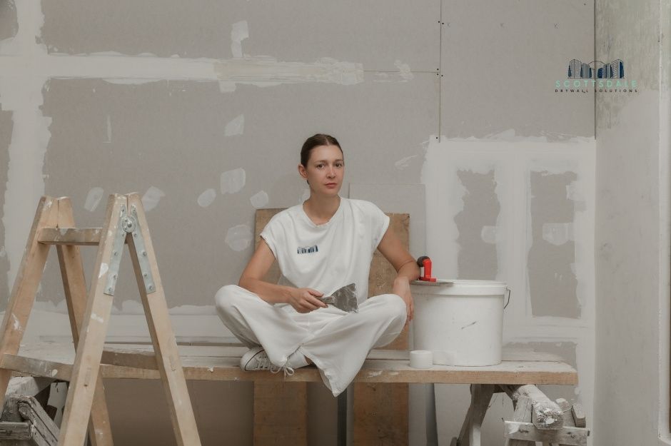 A drywall repair company professional sits on a work table wearing a white t-shirt and white pants in a room under residential construction. She holds a putty knife tool with white walls, plastic sheeting, and a ladder visible nearby near S Glenburn Dr, and E Cochise Pl, Sun Lakes, AZ.