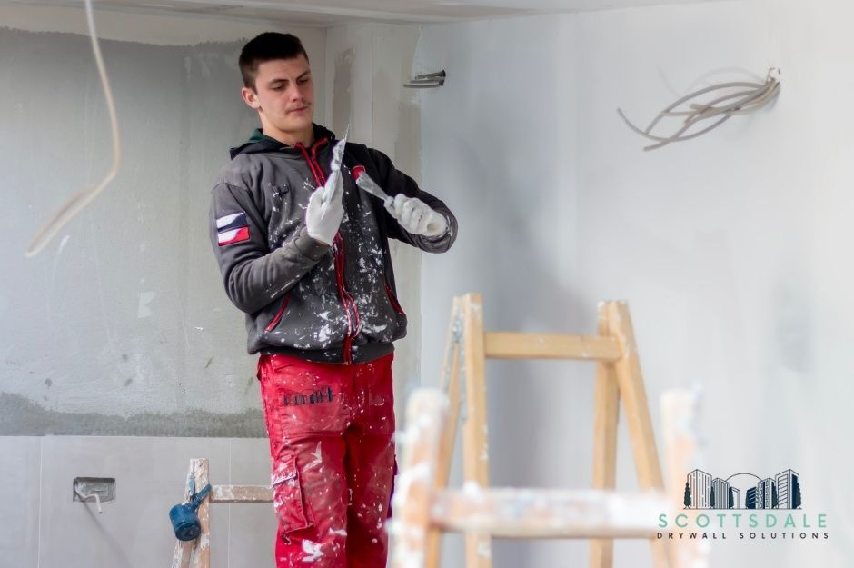 A drywall repair company worker at a residential construction site uses a drywall knife to apply joint compound on white walls. He wears paint-splattered red pants and a dark jacket while standing on wooden scaffolding near W Orchid Ln, and N 181st Ave, Waddell, AZ.
