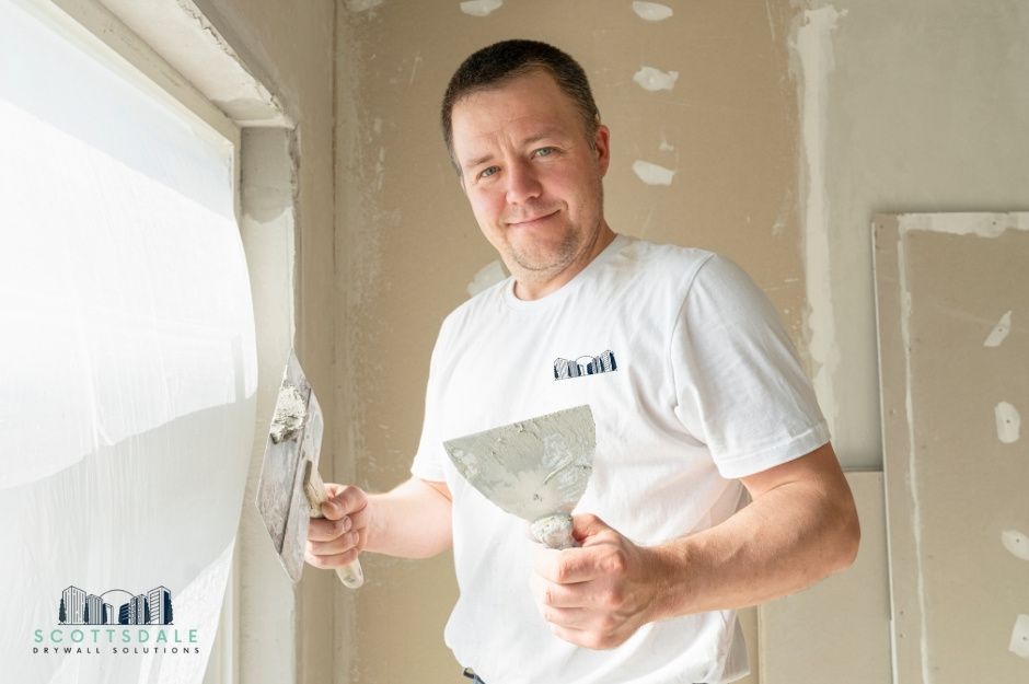 A drywall repair contractor in a white t-shirt smiles while holding a putty knife and joint compound. He stands inside a residential room under construction, where multiple circular patches show repair work in progress. Located near E Chuckwalla Trail, and N Canyon Ridge Trail, Cave Creek, AZ.