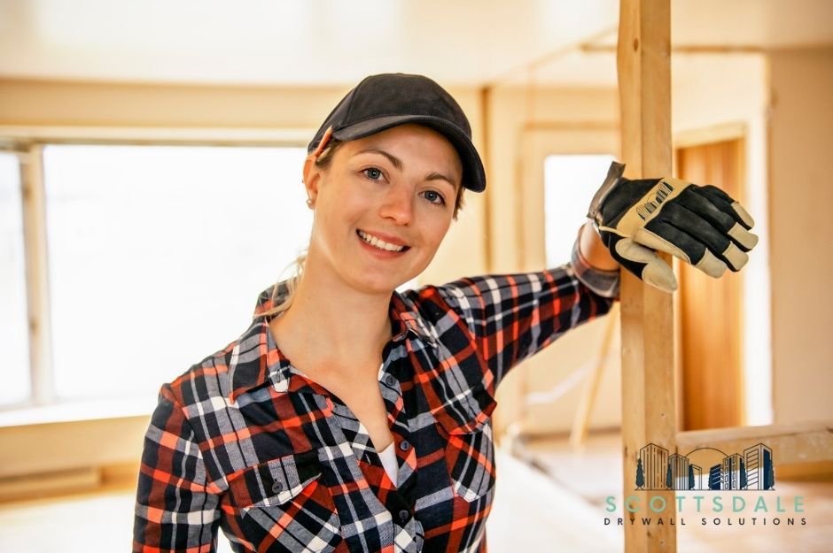 A drywall repair contractor wearing a black baseball cap, plaid flannel shirt, and work gloves stands smiling in a wood-framed residential construction site with exposed studs and drywall panels visible. Located near E Mesquite Cir, and Desierto Dr, Rio Verde, AZ.