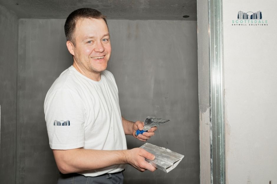 A drywall repair contractor smiles while holding a putty knife and sanding block, working on smoothing joint compound on a wall in an unfinished room with exposed brick and metal framing during a residential renovation project near S El Camino Dr, and E Encanto Dr, Tempe, AZ.