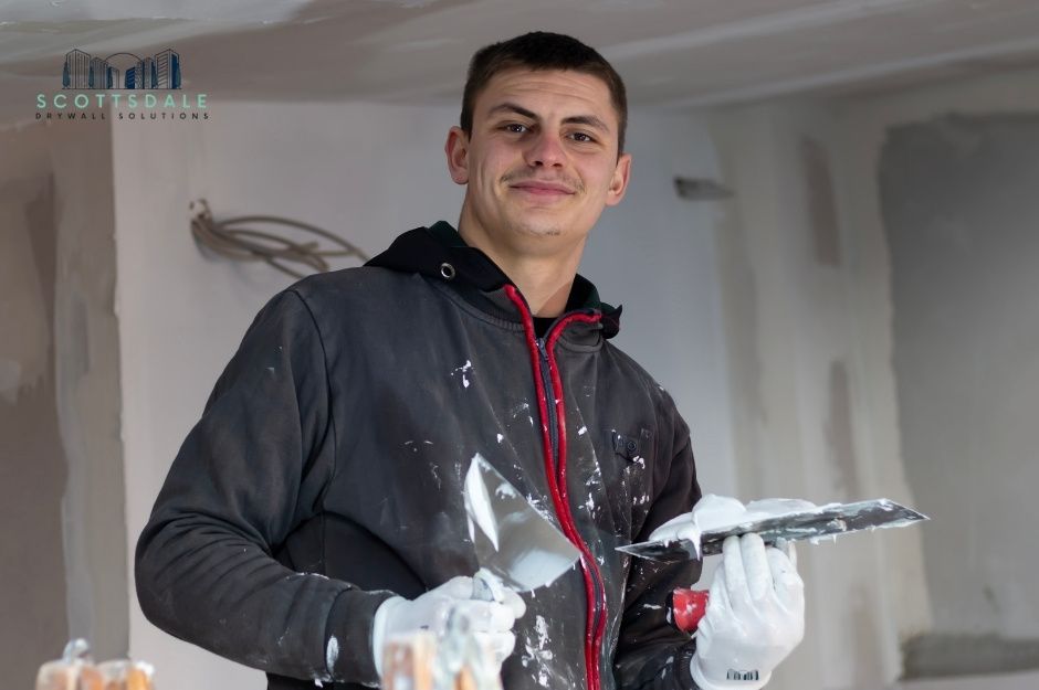 A drywall repair handyman smiling while holding a putty knife and trowel, wearing white gloves and a black hoodie with red drawstrings. He is covered in white drywall compound splatters from repair work at a residential construction site near W Marshall Ave, and W Missouri Ave, Citrus Park, AZ.