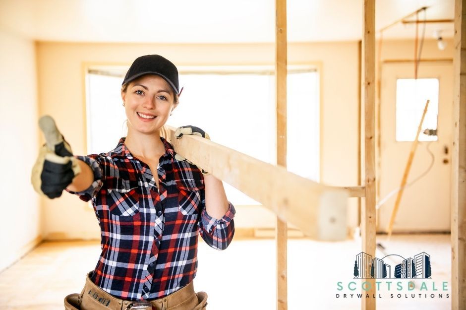 A smiling female drywall repair technician wearing a black cap and red plaid shirt carries a wood over her shoulder in an unfinished residential construction site with exposed wood framing and concrete floors, located near S Griswold St, and E Ginko Ct, Gilbert, AZ.