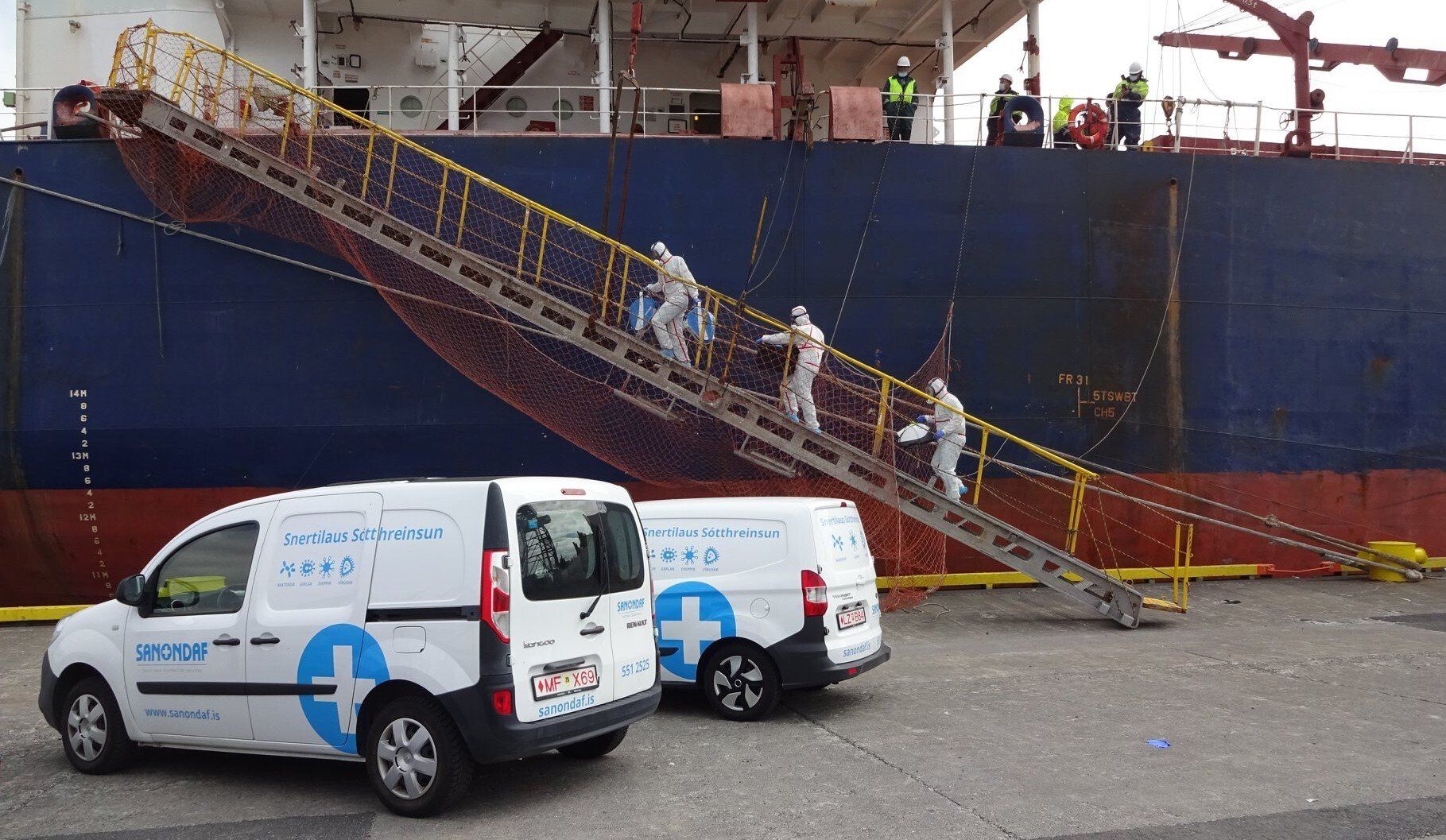 People in hazmat suits climbing gangway onto a blue ship, parked near two vans with medical logos.