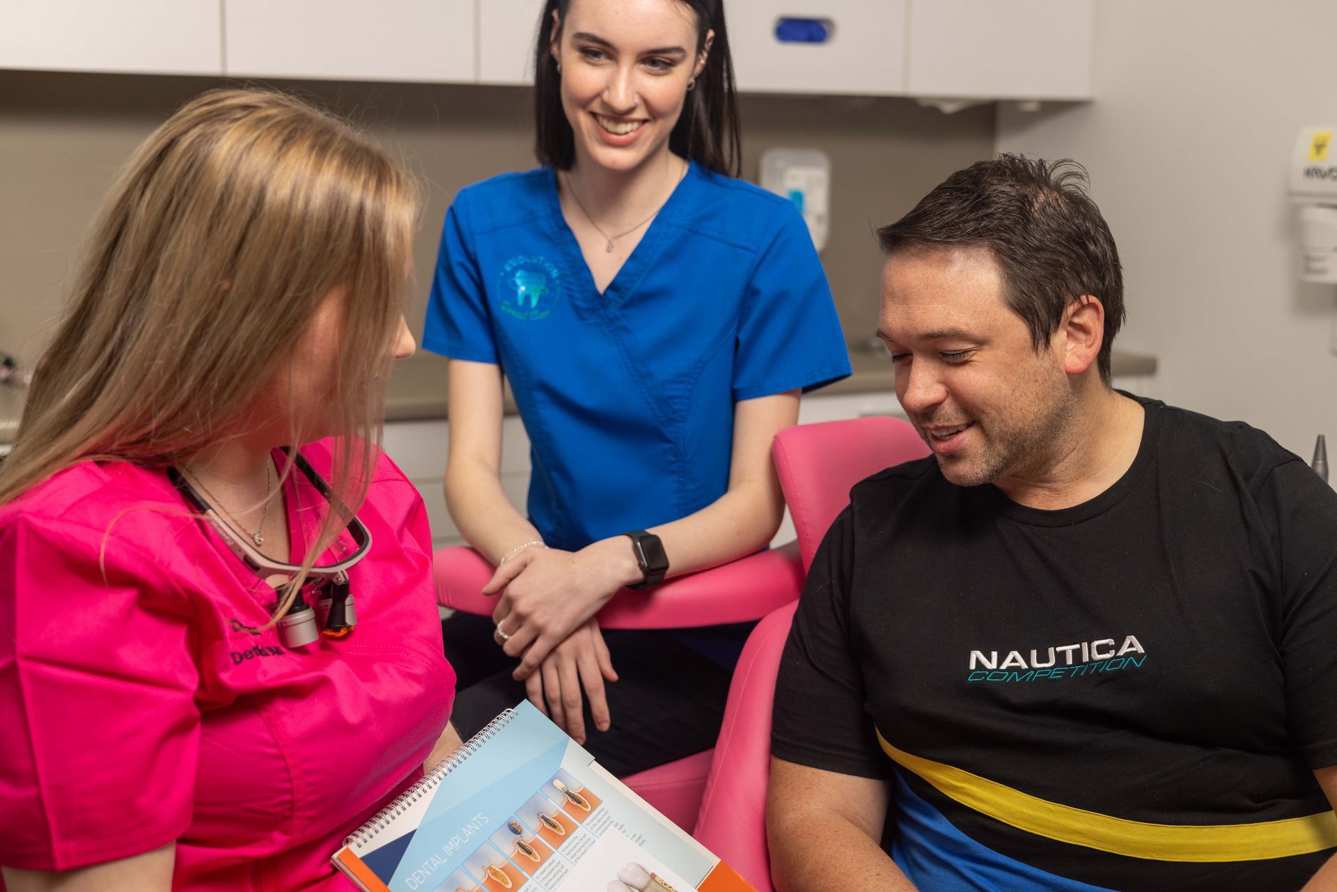 A man is sitting in a dental chair talking to two nurses.