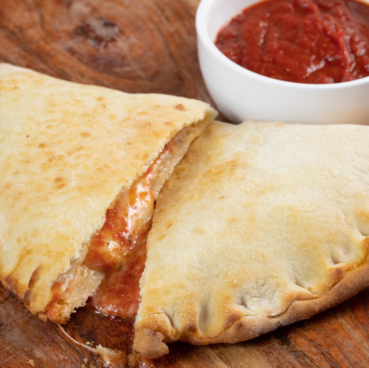 A calzone is sitting on a wooden table next to a bowl of sauce.