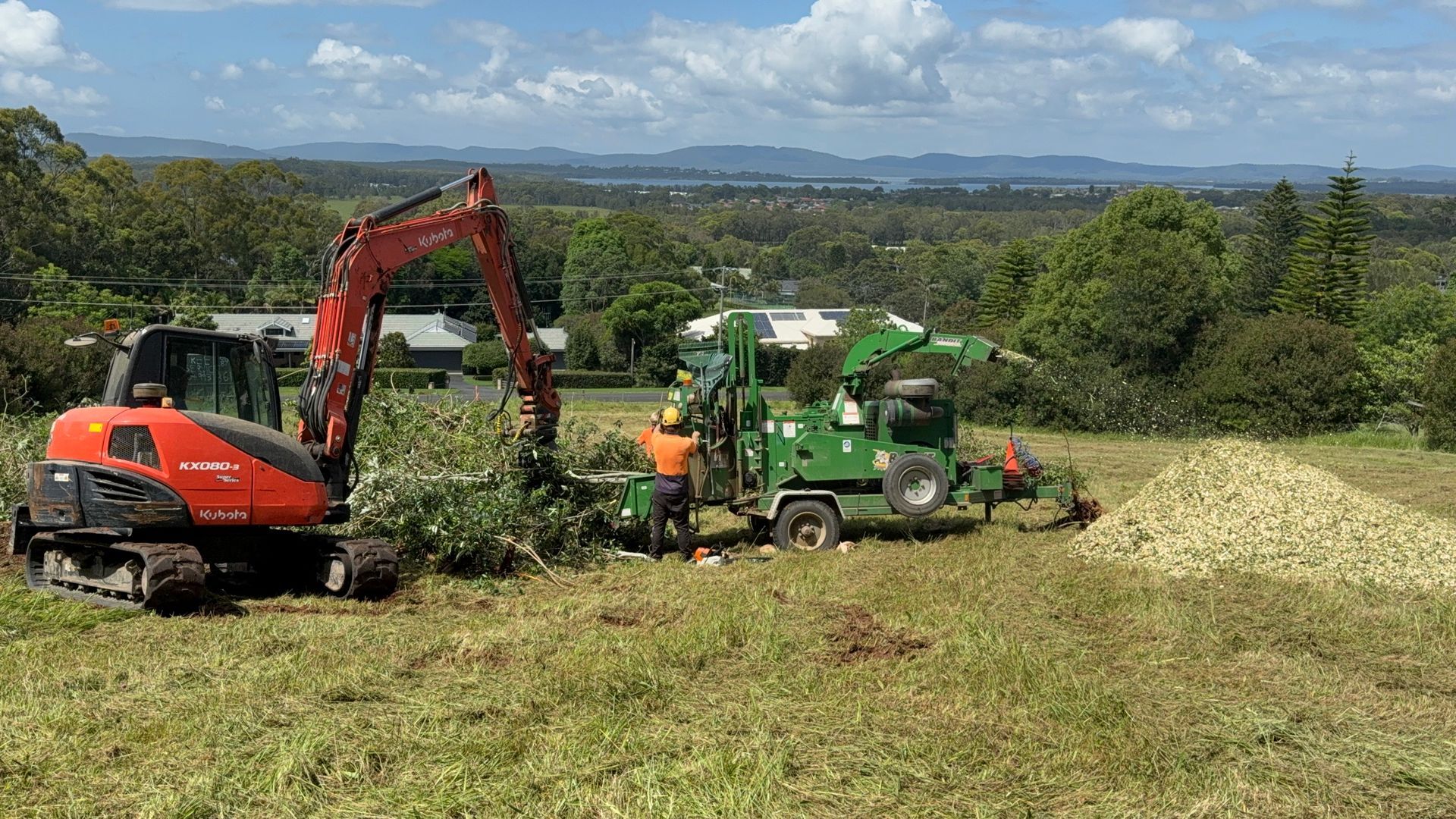 An excavator feeds branches into a wood chipper; a worker monitors — Coastal Slashing & Mowing Services In Harrington, NSW