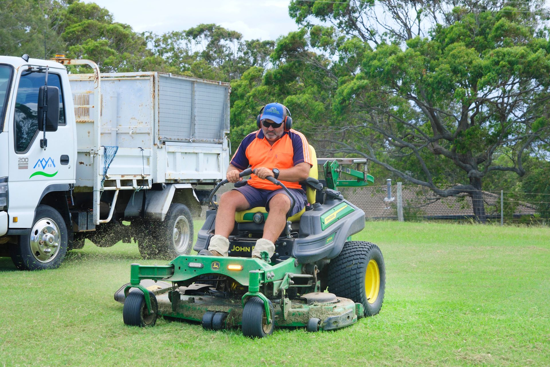 A person wearing a high-visibility orange shirt and hearing protection mows a lawn on a green John Deere ride-on mower. — Coastal Slashing & Mowing Services In Tallwoods Village, NSW