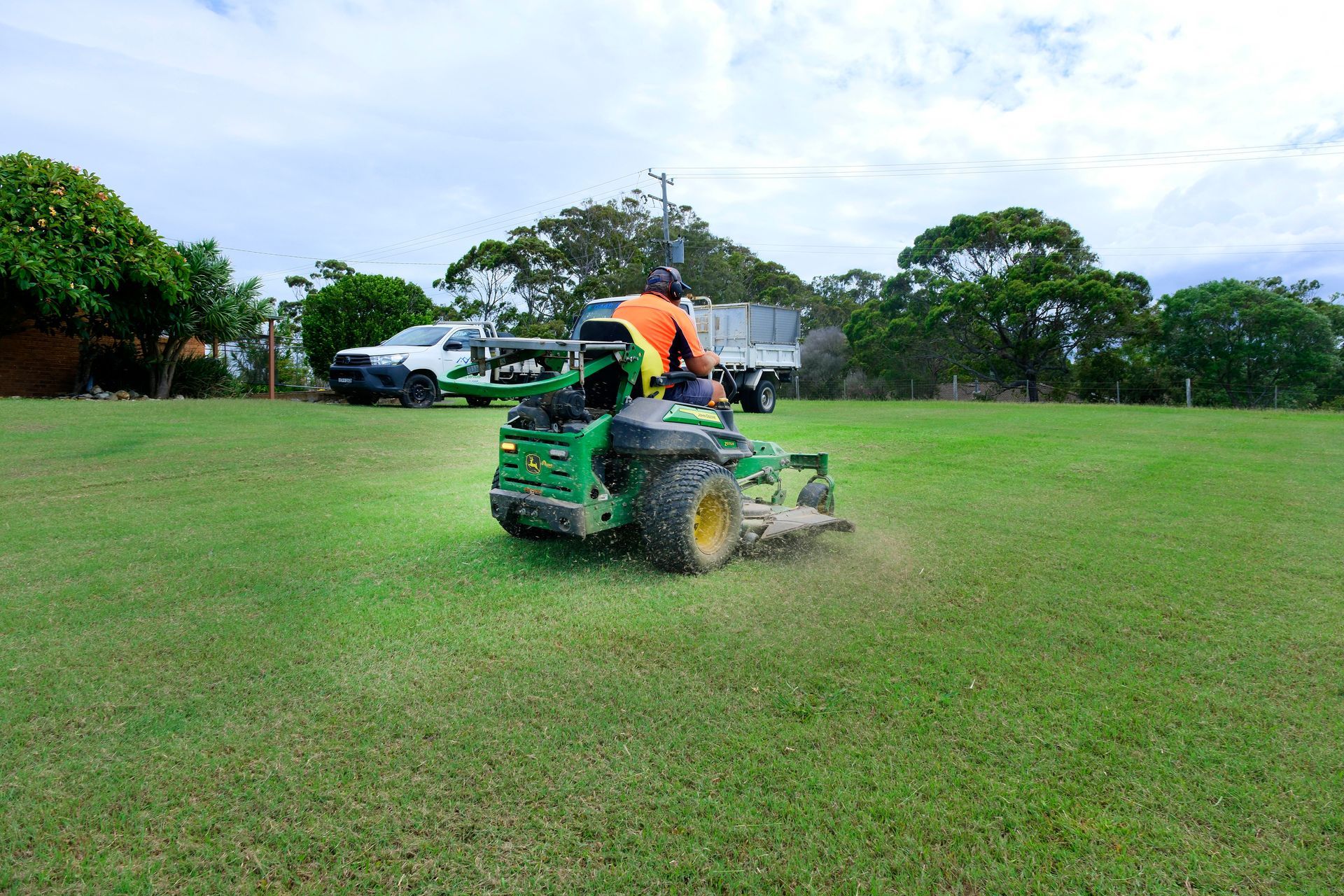 A Person in an Orange Shirt Mows a Large, Green Grassy Field — Coastal Slashing & Mowing Services In Tallwoods Village, NSW