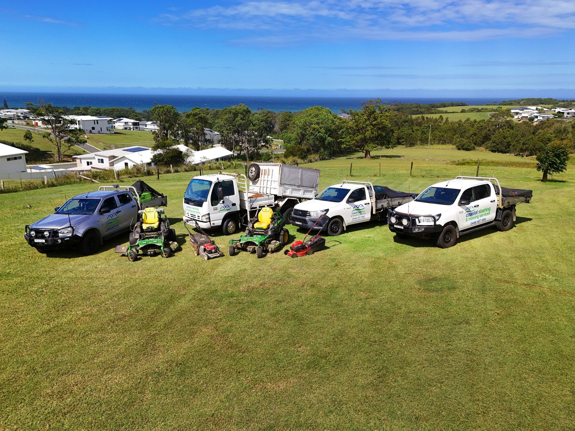 Three white utility trucks and a silver SUV parked in a line on a grassy hill, with lawn equipment and the ocean behind. — Coastal Slashing & Mowing Services In Taree, NSW