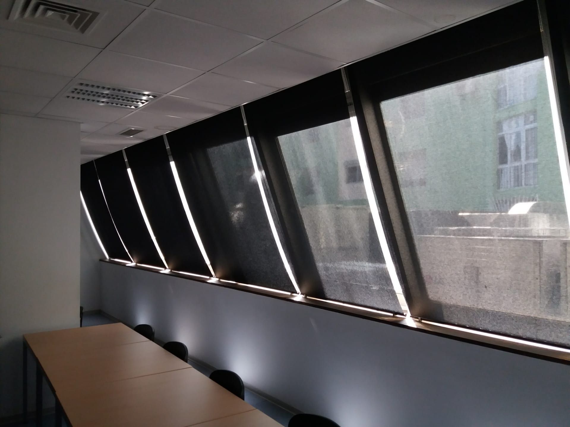 Sala de conferências com janelas angulares cobertas por persianas escuras; a luz do sol entra abundantemente.