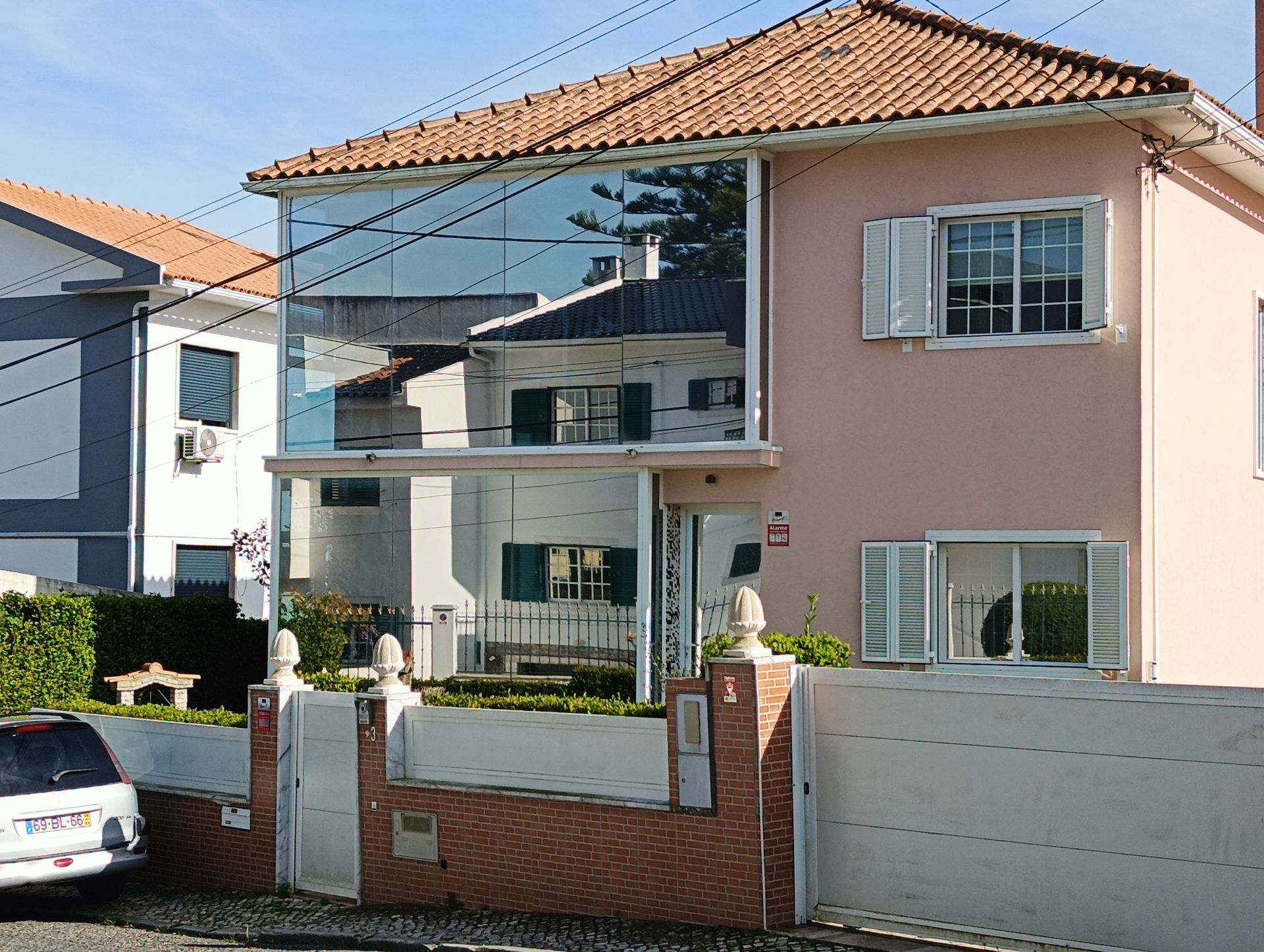Casa rosa de dois andares com seção espelhada que reflete as construções vizinhas. Persianas brancas, telhado de telha e parede de tijolos.