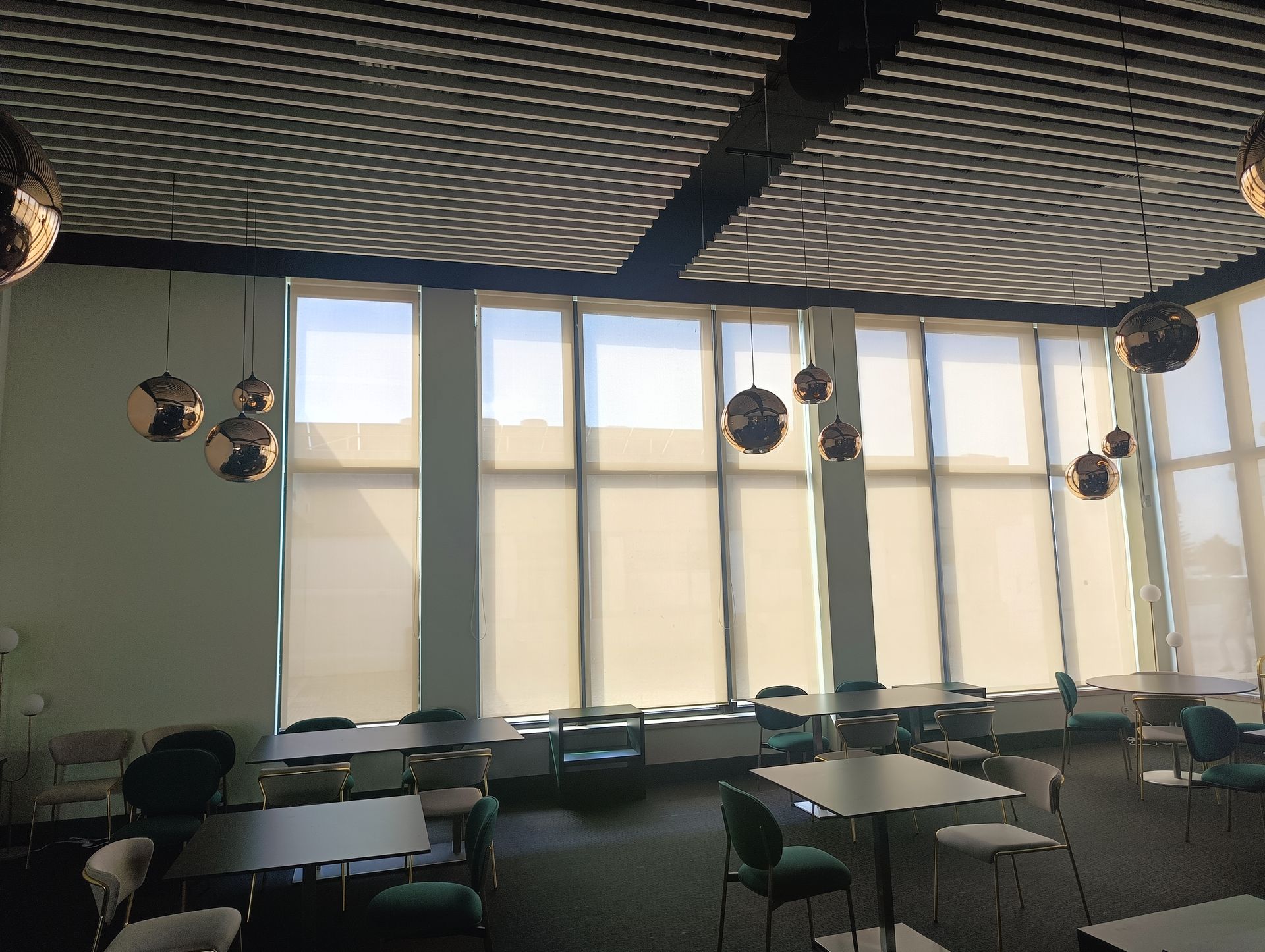 Uma sala ensolarada com mesas, cadeiras e grandes janelas com persianas. Luminárias decorativas em forma de globo pendem do teto.