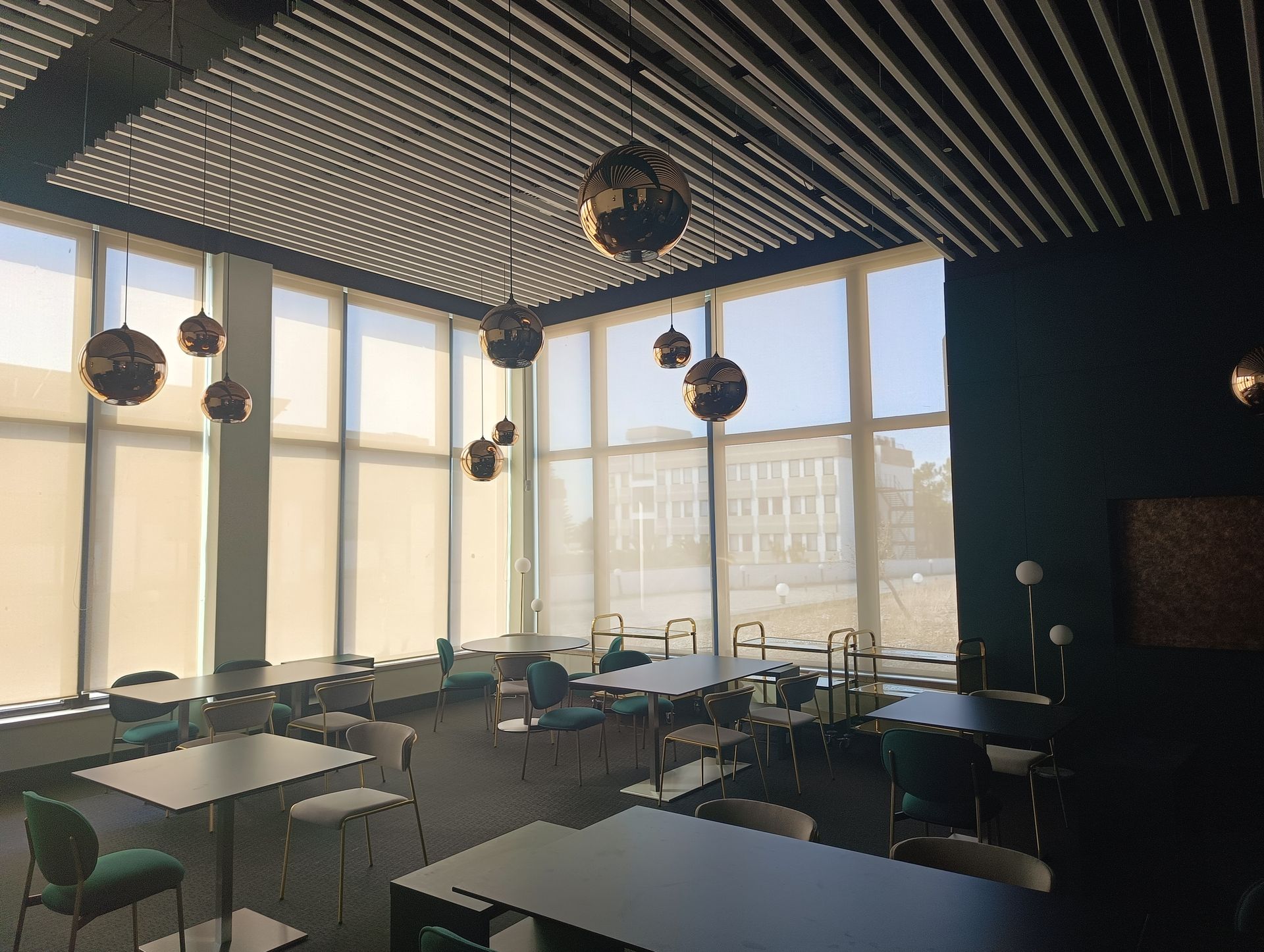 Interior de café com mesas, cadeiras, janelas amplas, luminárias pendentes em formato de globo e uma parede verde escura.