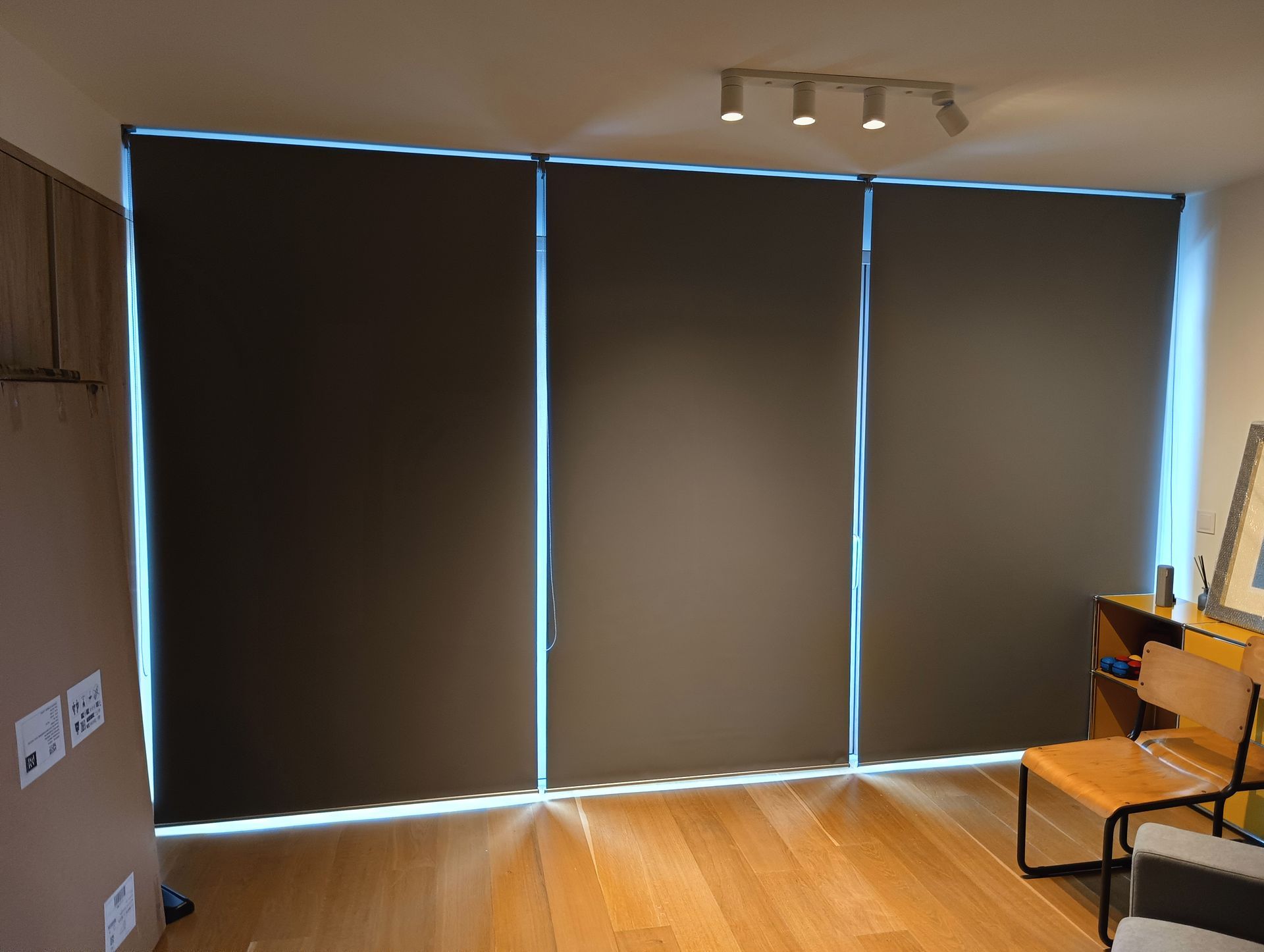 Três persianas de rolo cinza-escuras cobrem uma janela; uma luz brilha ao redor das bordas.