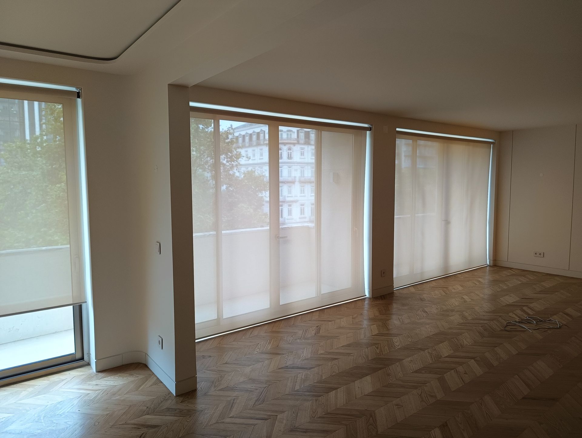 Quarto vazio com cortinas bege cobrindo as janelas, piso de parquet e paredes brancas.