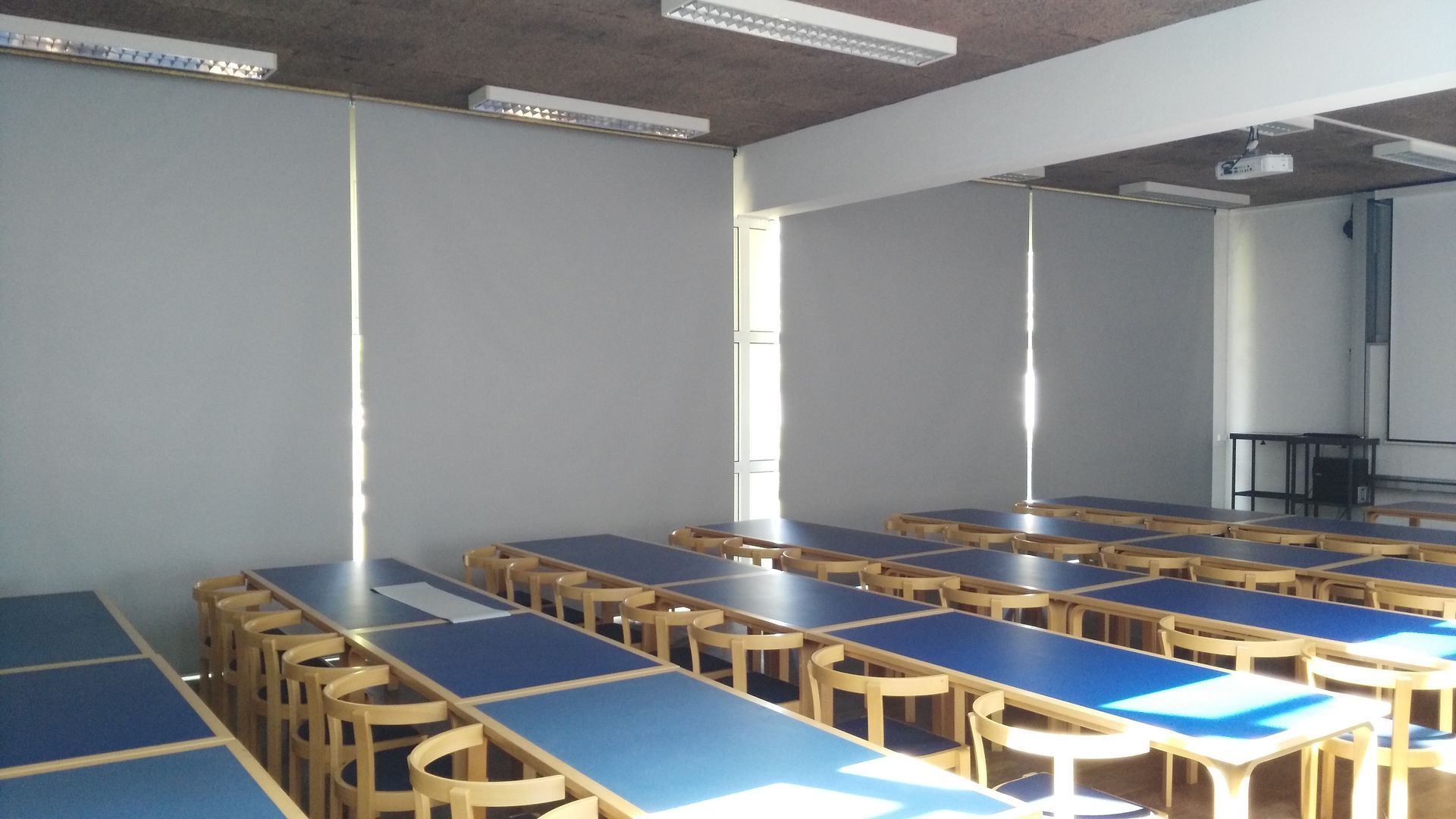 Fileiras de mesas com tampo azul e bancos de madeira em uma sala de aula com paredes brancas, janela e luzes.