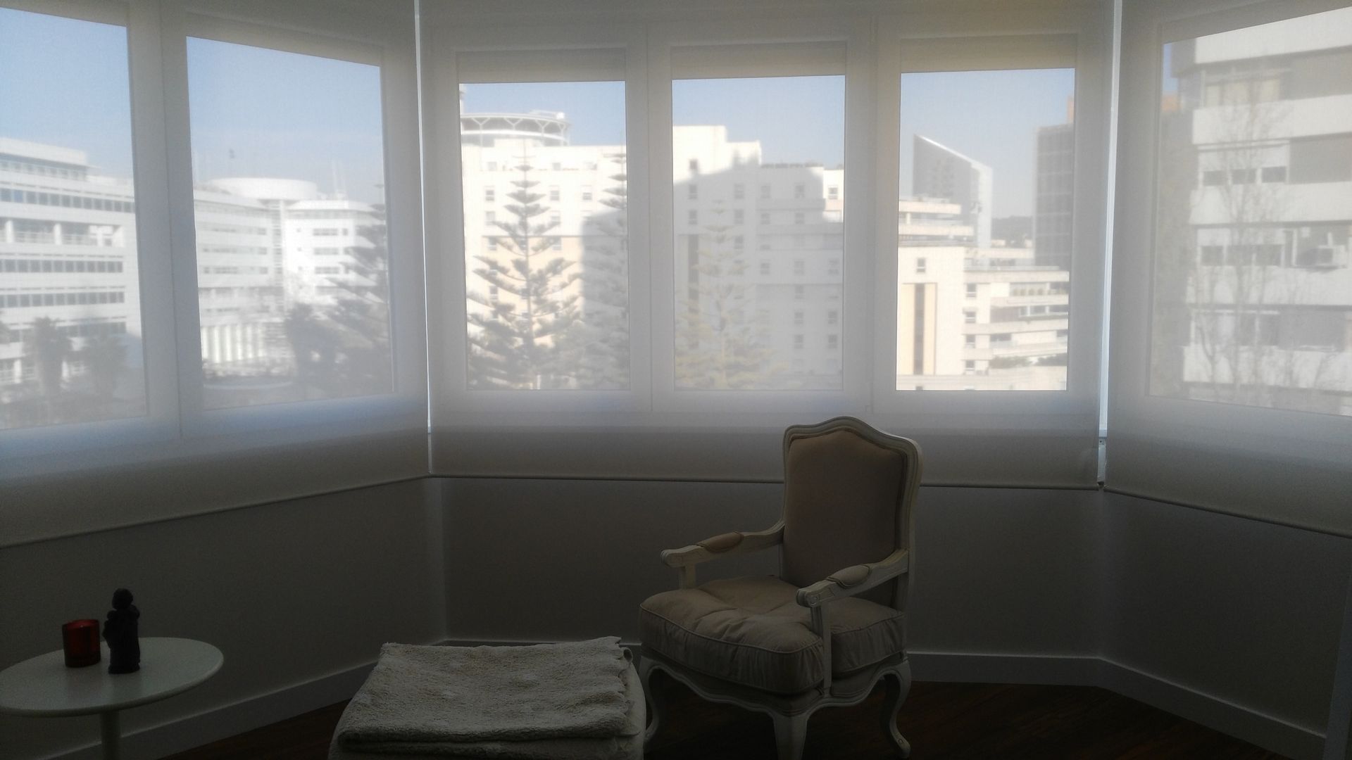 Um quarto com persianas brancas, uma cadeira estampada e vista para a cidade.