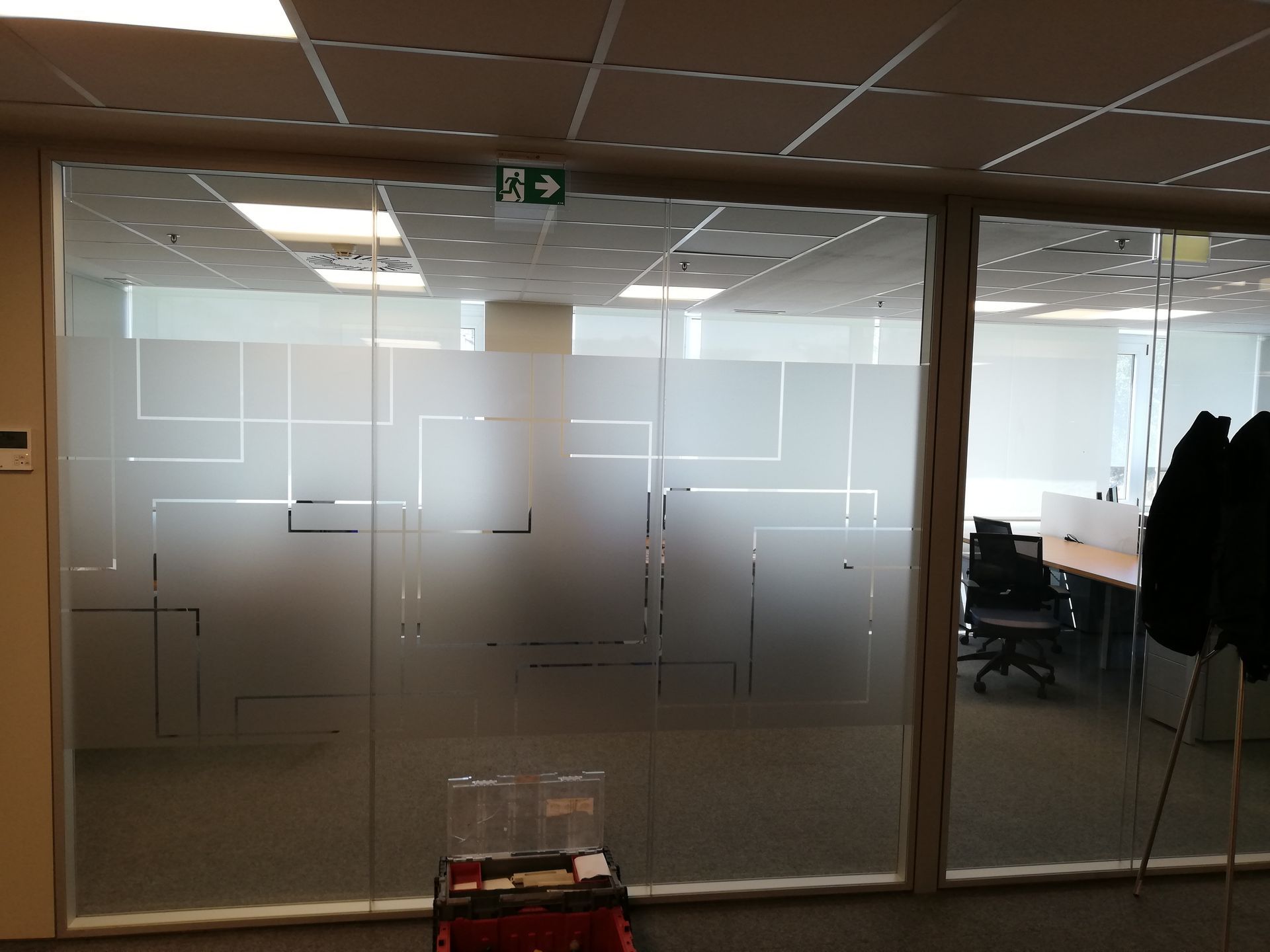 Escritório com paredes de vidro fosco, desenhos geométricos e placa de saída de emergência.