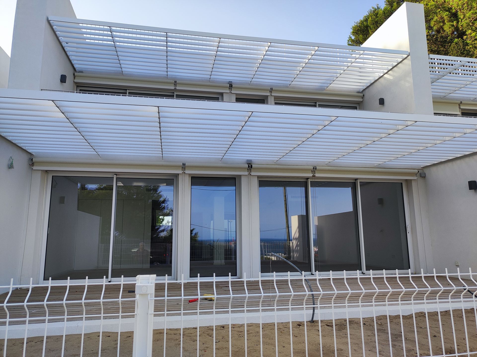 Prédio branco com grandes janelas e telhado em treliça; uma cerca branca está na frente.