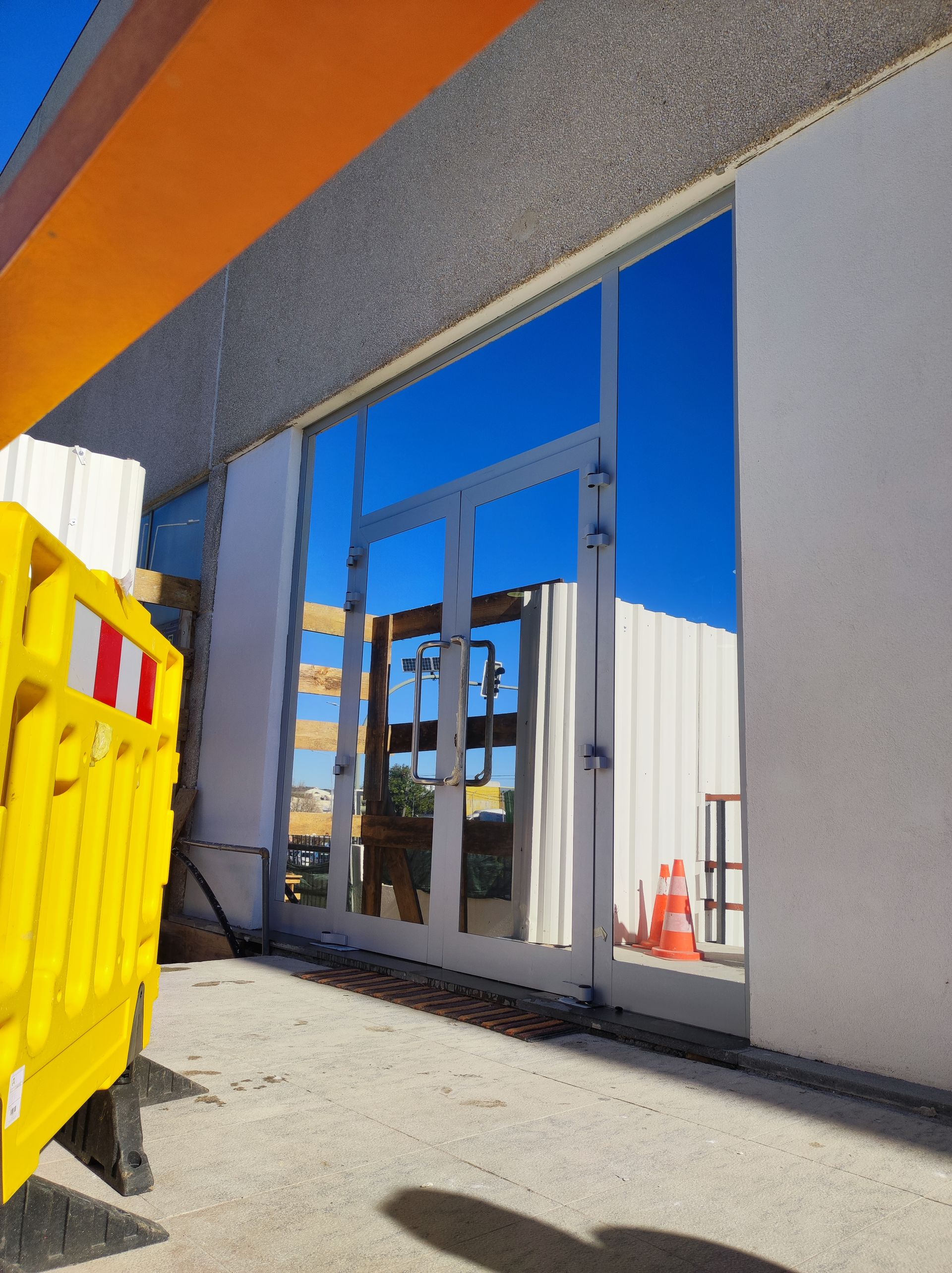 Porta de vidro refletindo o céu azul, ladeada por paredes brancas e uma barreira de construção laranja.