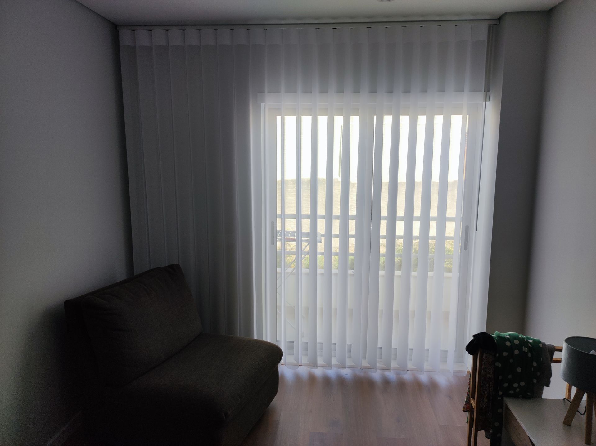 Quarto com cortinas brancas translúcidas sobre uma porta/janela, um sofá marrom e uma mesa escura.