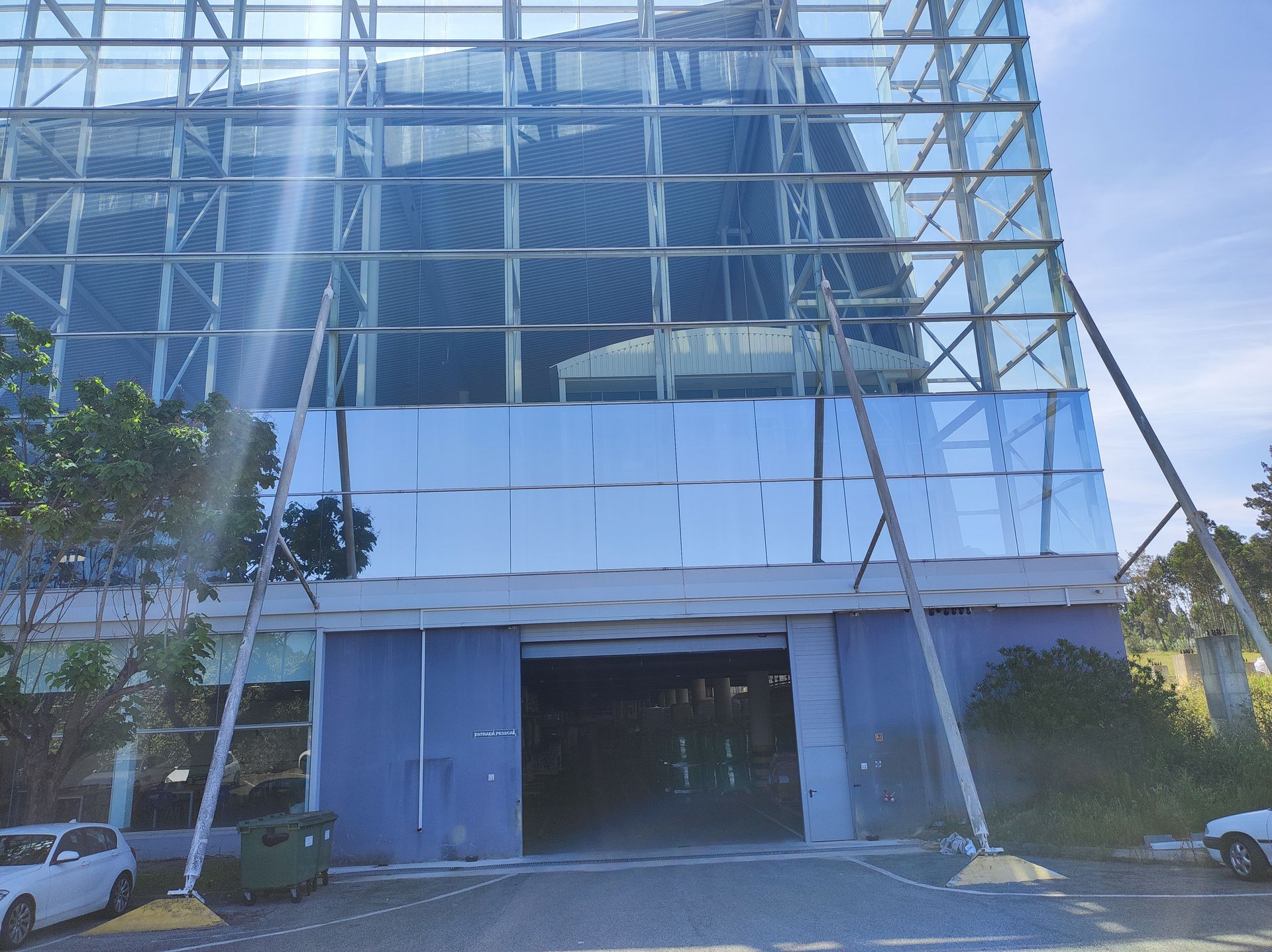 Edifício com fachada de vidro, sustentado por vigas metálicas. Painéis azuis e entrada aberta na base.