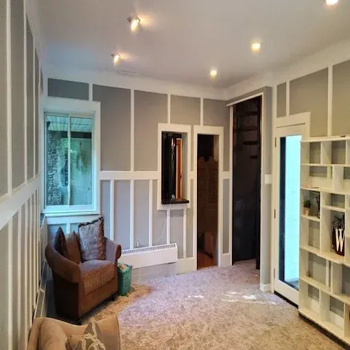 Cozy, gray-walled room with an armchair, built-in shelving, and a window overlooking greenery.