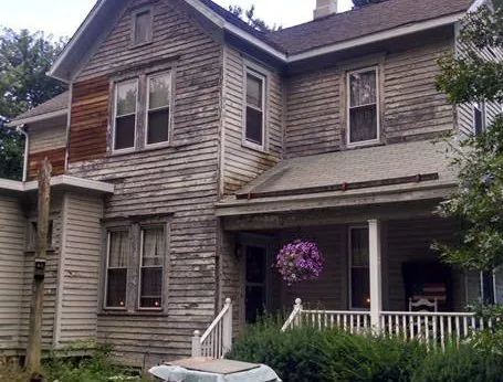 Old, weathered two-story house with peeling paint, porch, and overgrown bushes.