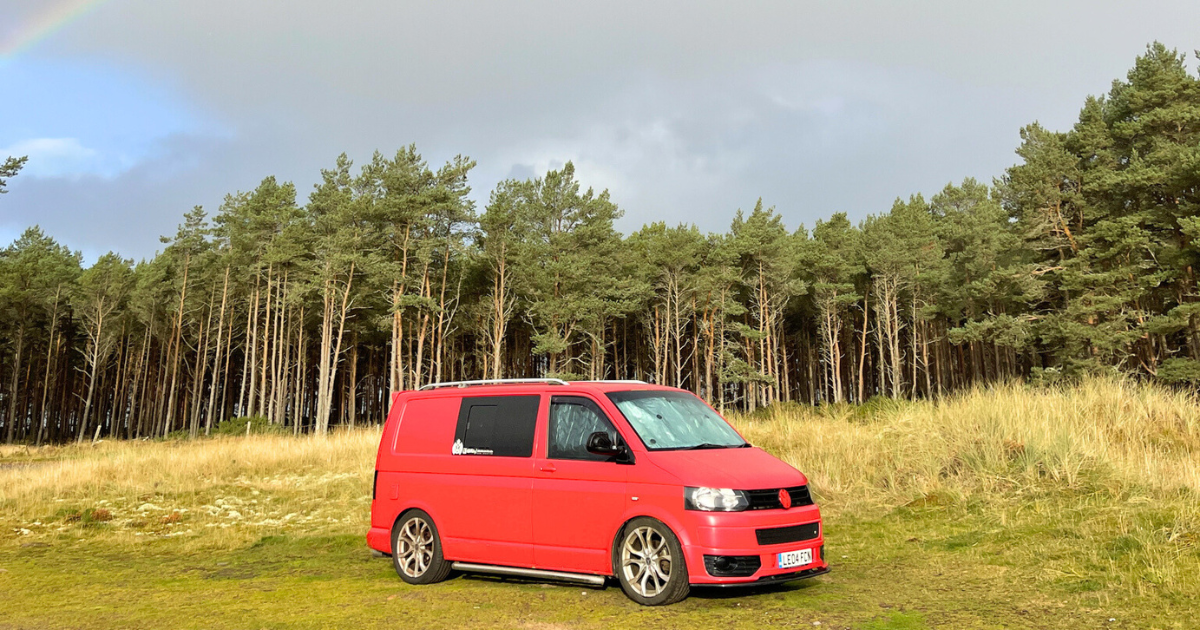 A red van is parked in a field with a rainbow in the background.