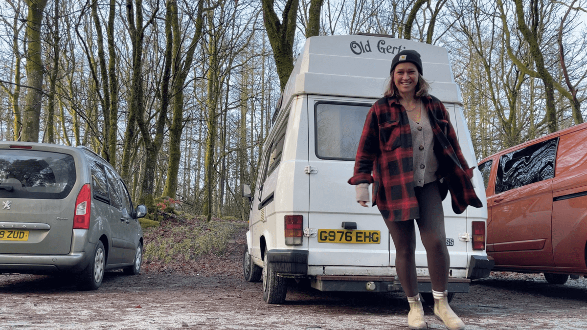 Kez is standing in front of a white campervan in a car park.