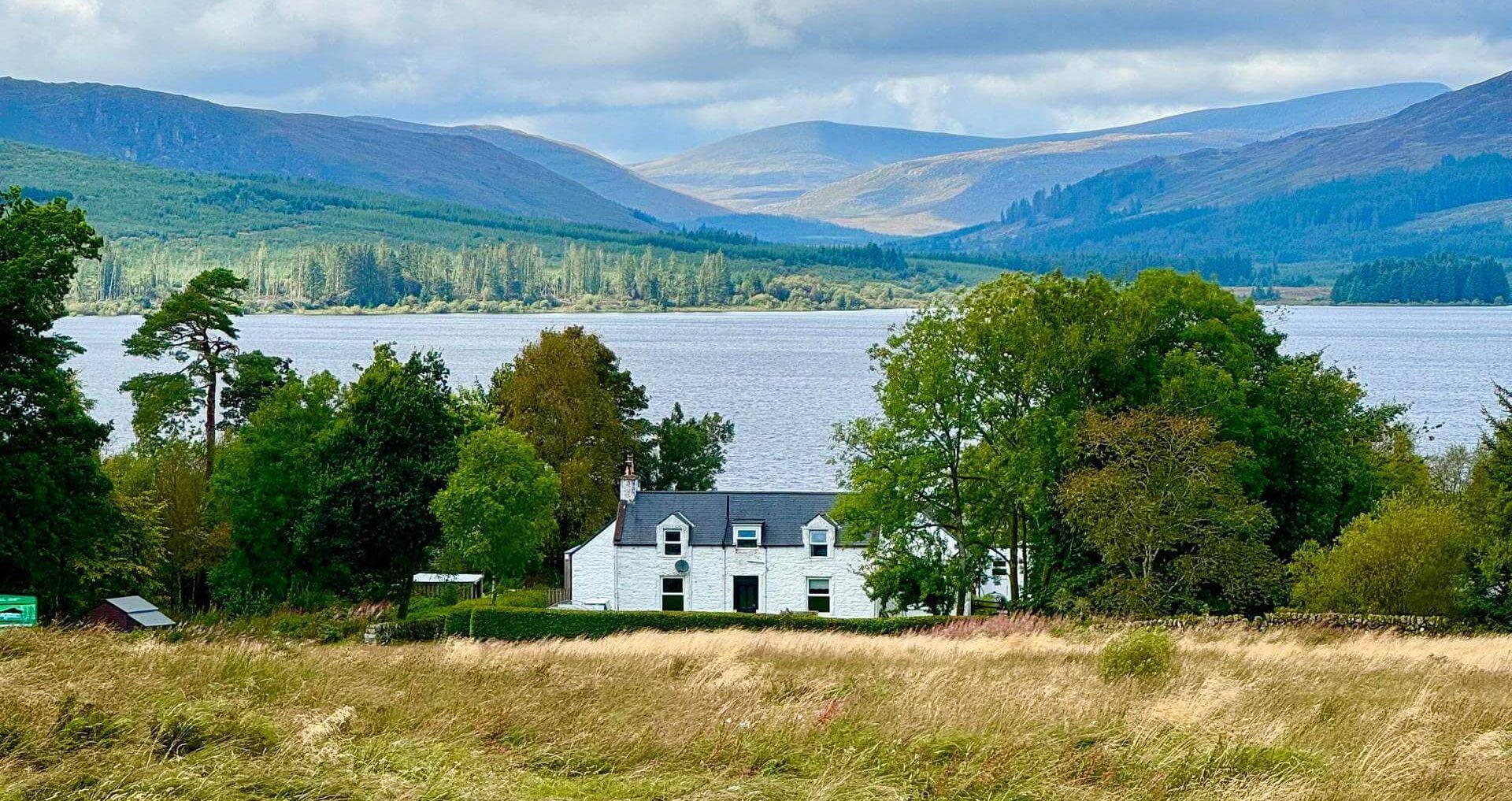There is a house in the middle of a field with a lake in the background.