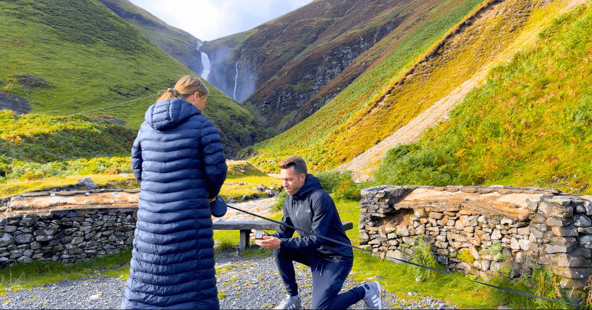 A man is kneeling down to propose to a woman in front of a mountain.