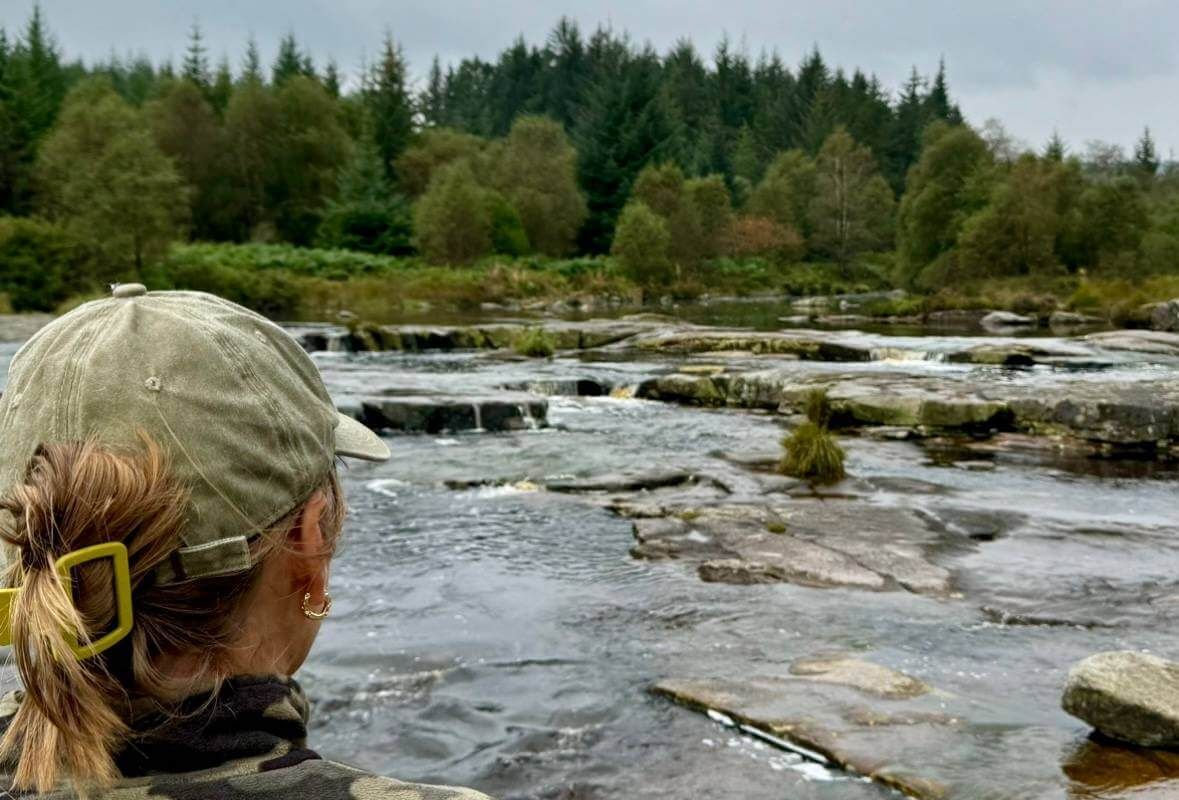 A woman in a baseball cap is looking at a river.