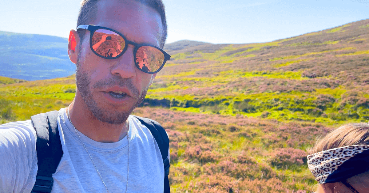 A man wearing sunglasses and a backpack is taking a selfie in a field.