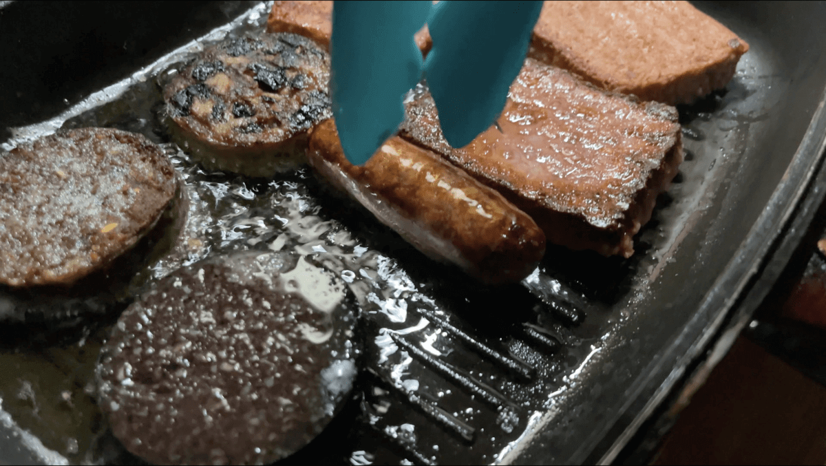 A pan filled with meat and sausages is being stirred with tongs.