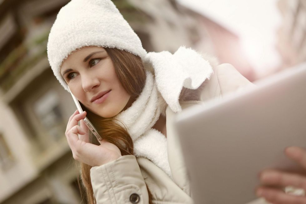 A woman wearing a white hat and scarf is talking on a cell phone while looking at a tablet.