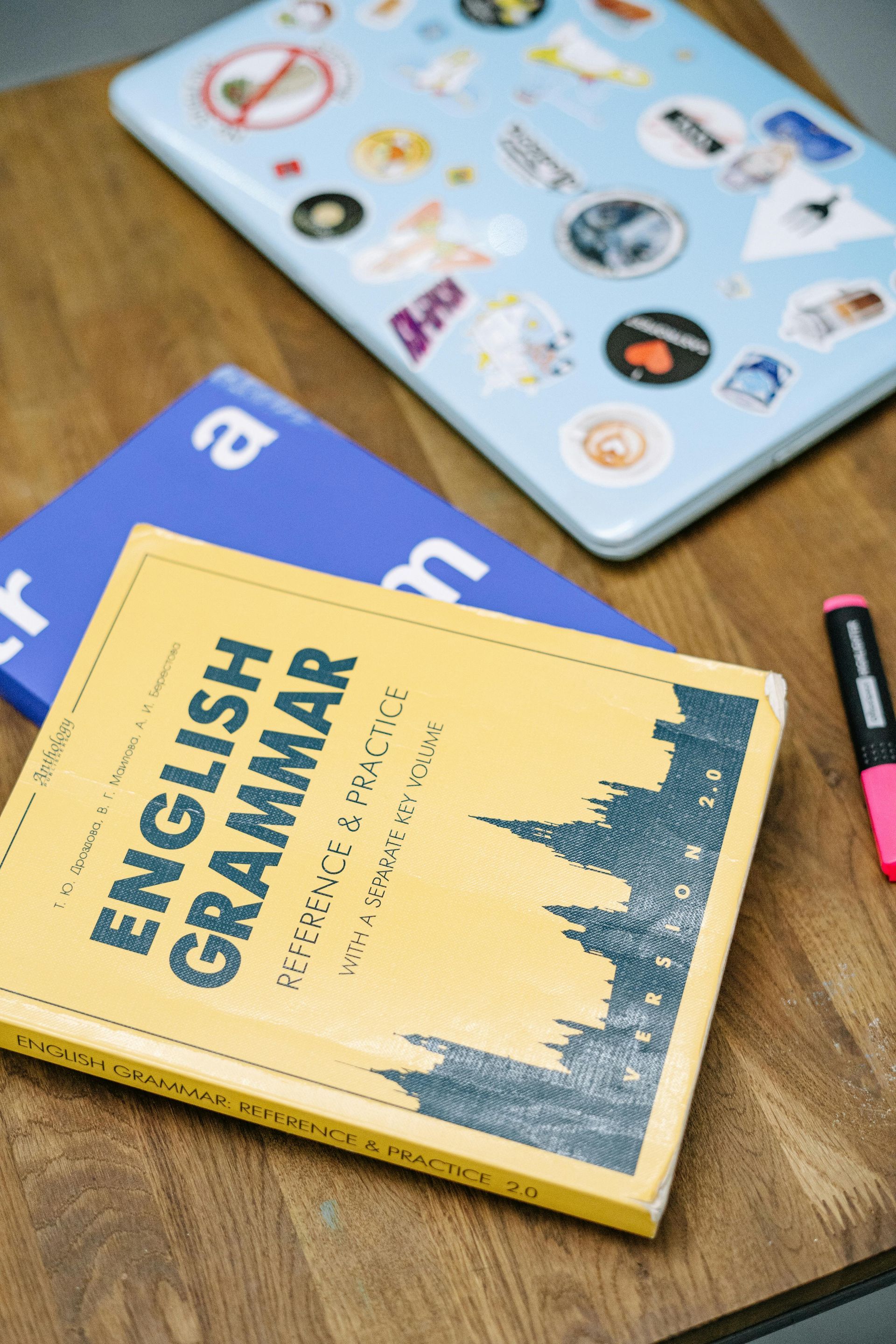 A book titled english grammar is sitting on a wooden table next to a laptop.