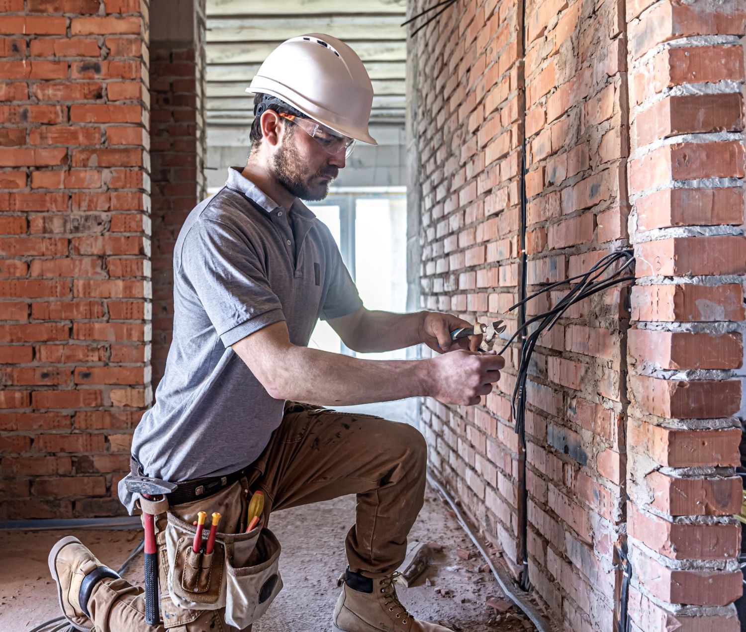 Person installing electrical wiring on a brick wall, using a ladder. Red conduits run across the ceiling and wall.