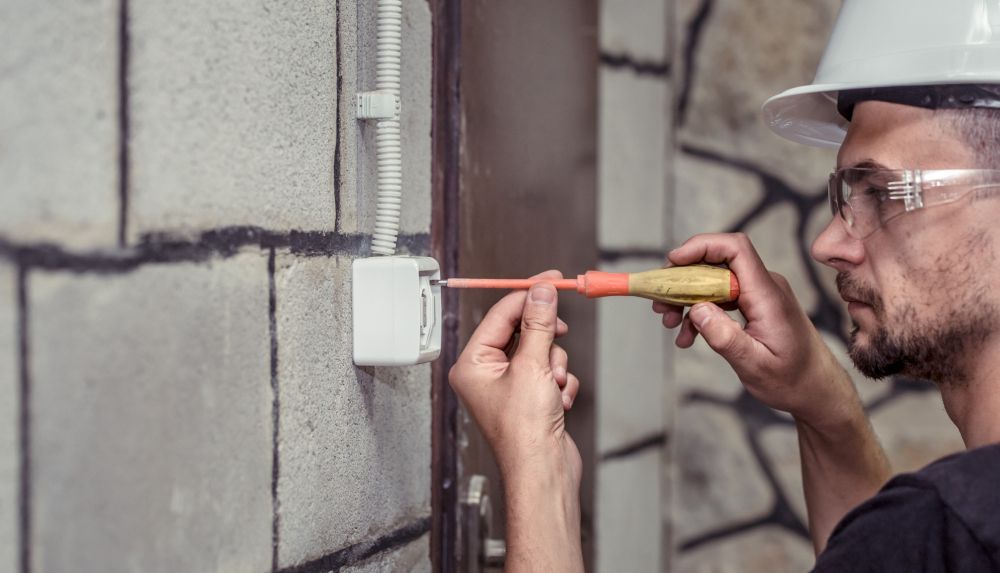 Hand with screwdriver connecting wires to electrical outlet on white wall.