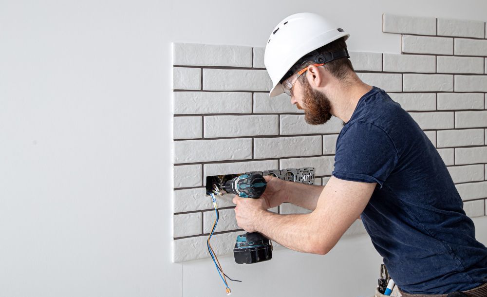Electrician's hand holding a screwdriver, working on a double electrical outlet installed in a white wall.