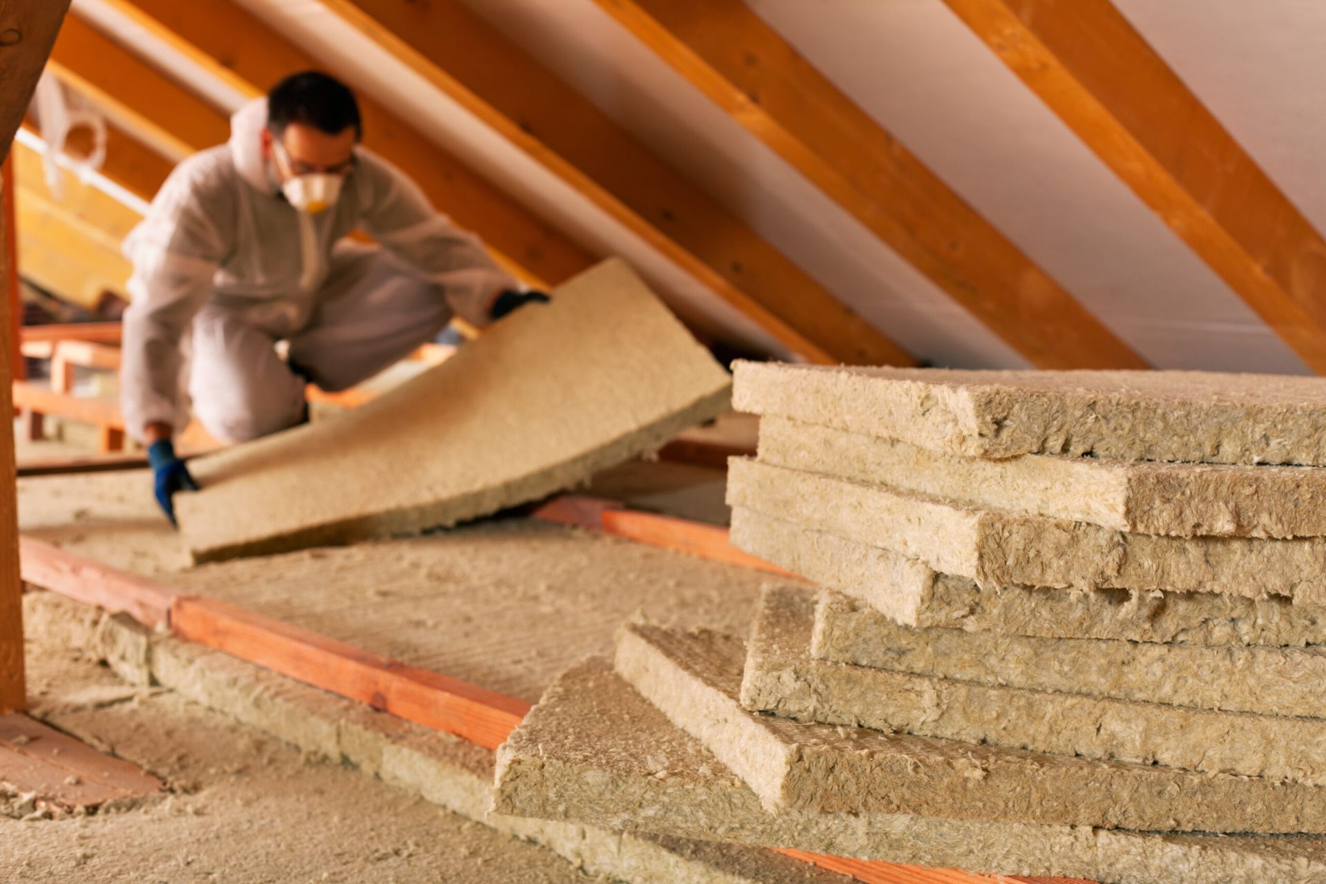 Person installing insulation in an attic. Beige insulation, wooden beams, worker in protective gear.