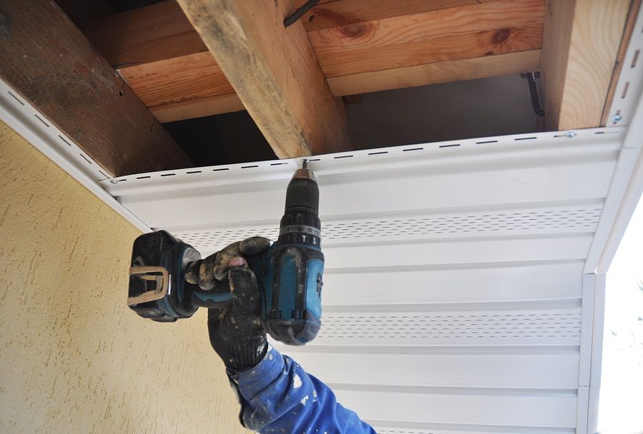 Person using a drill to install white vinyl siding under a wooden beam.