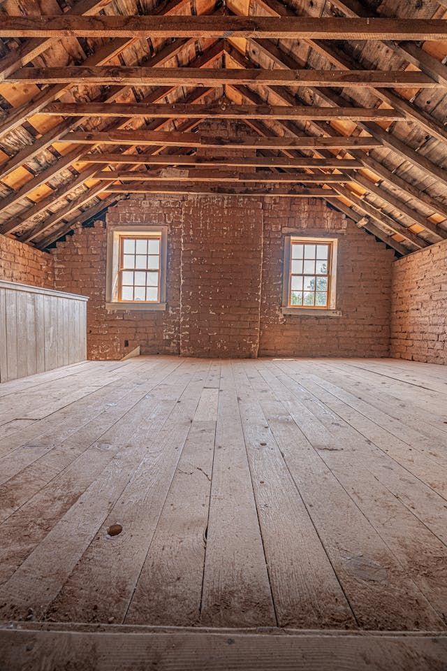Empty attic with wooden floor, exposed beams, brick walls, two windows, and natural light.