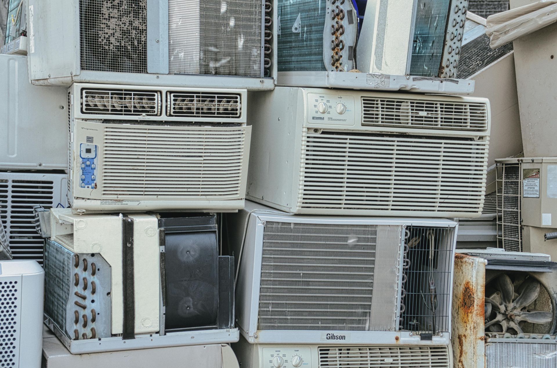 Pile of stacked, used window air conditioners; mostly white, some with rust and wear.