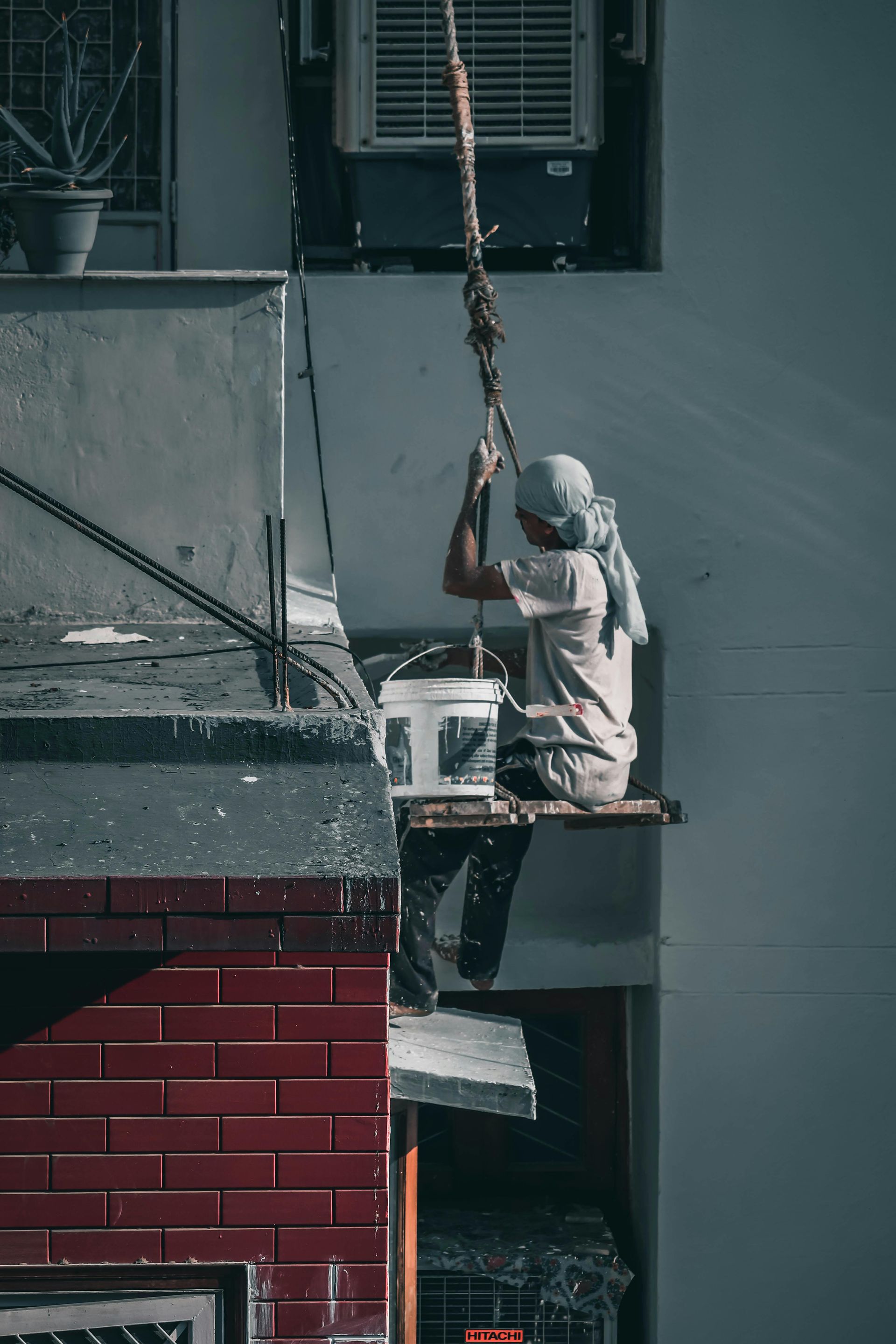 Person on a precarious platform, pulling a bucket suspended from a rope. Beside a building with a red brick wall.