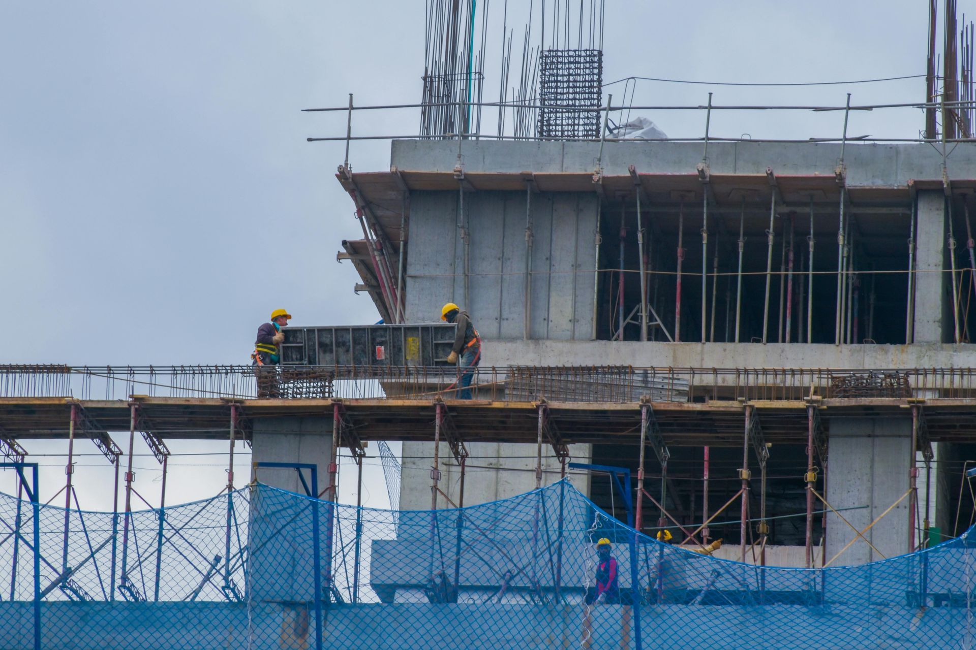Construction workers on scaffolding, setting up forms at a building site with blue safety netting. Cloudy sky.