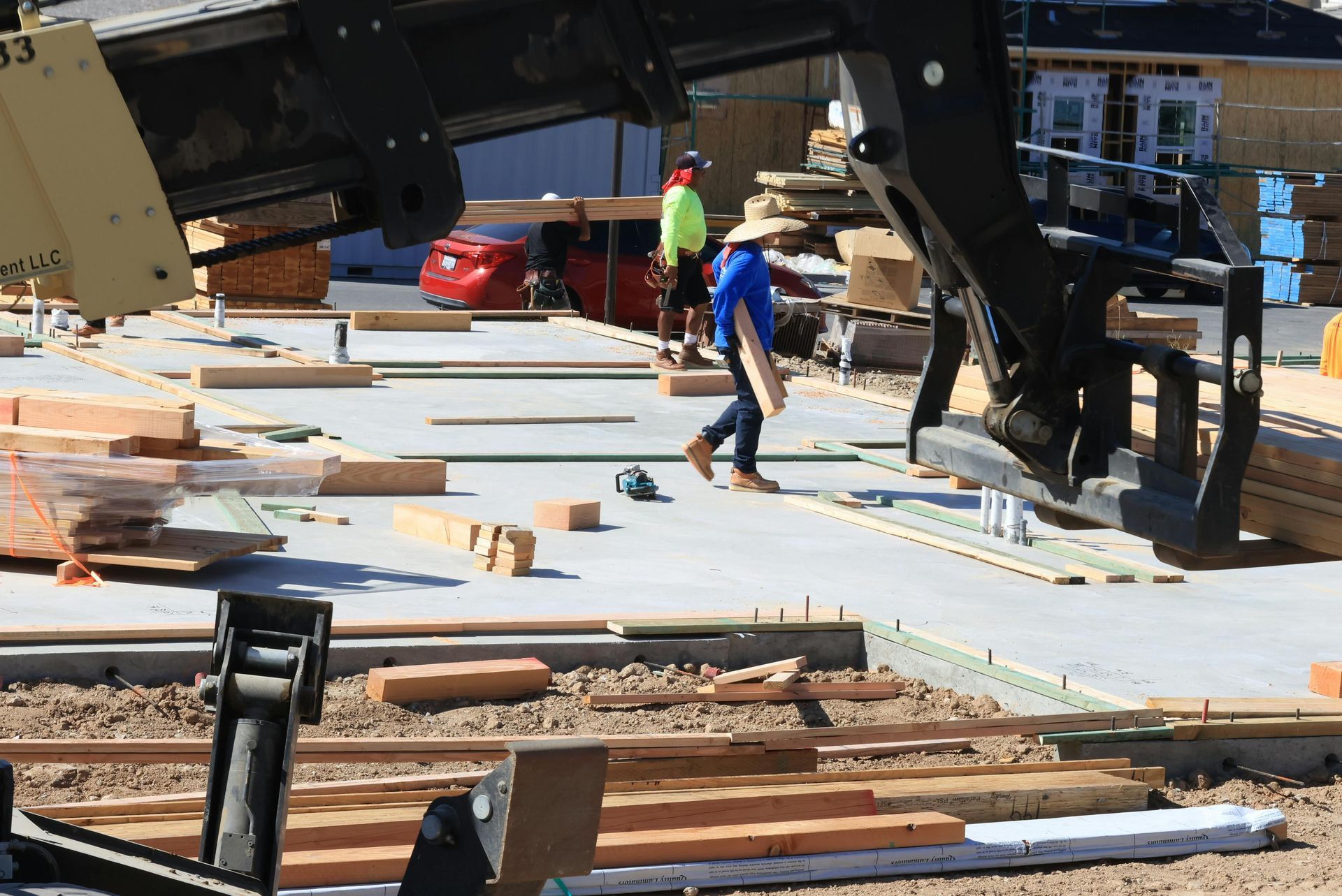 Construction site; worker carrying wood, excavator overhead. Concrete foundation, lumber, tools, a red car.