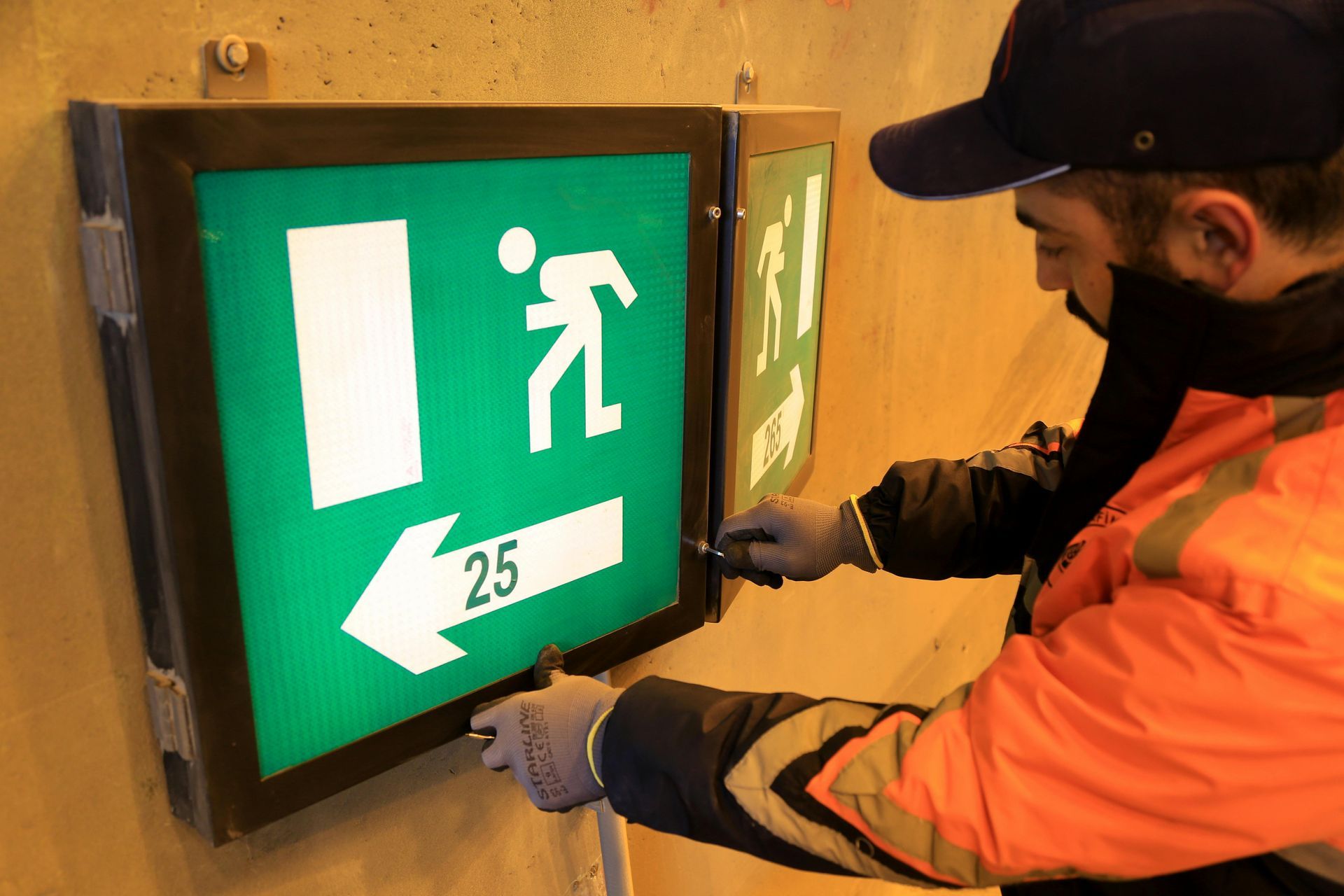 Person installing emergency exit sign, green with running person icon, left arrow, and number 25.