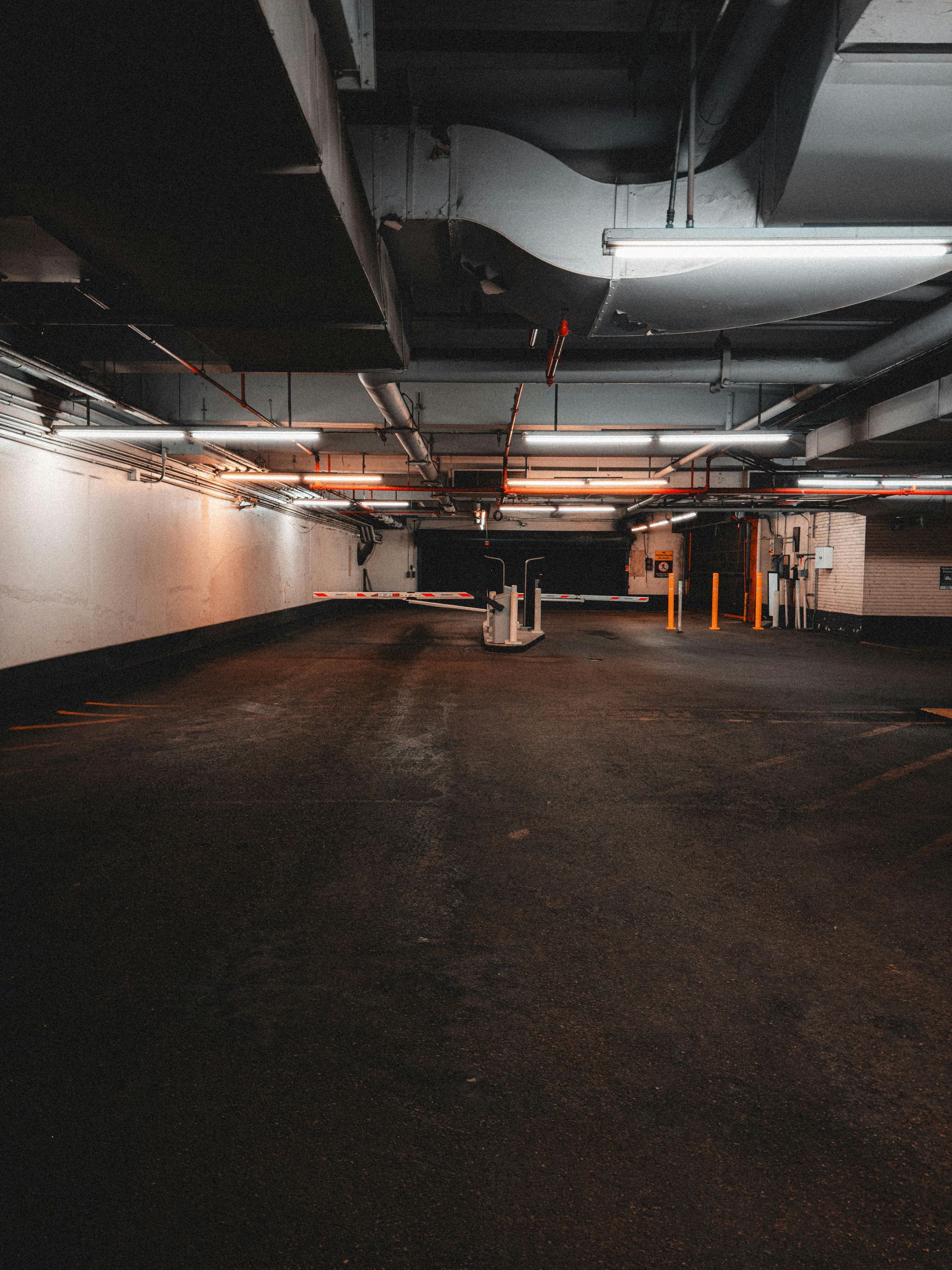 An empty, dimly lit parking garage with overhead pipes and fluorescent lights.